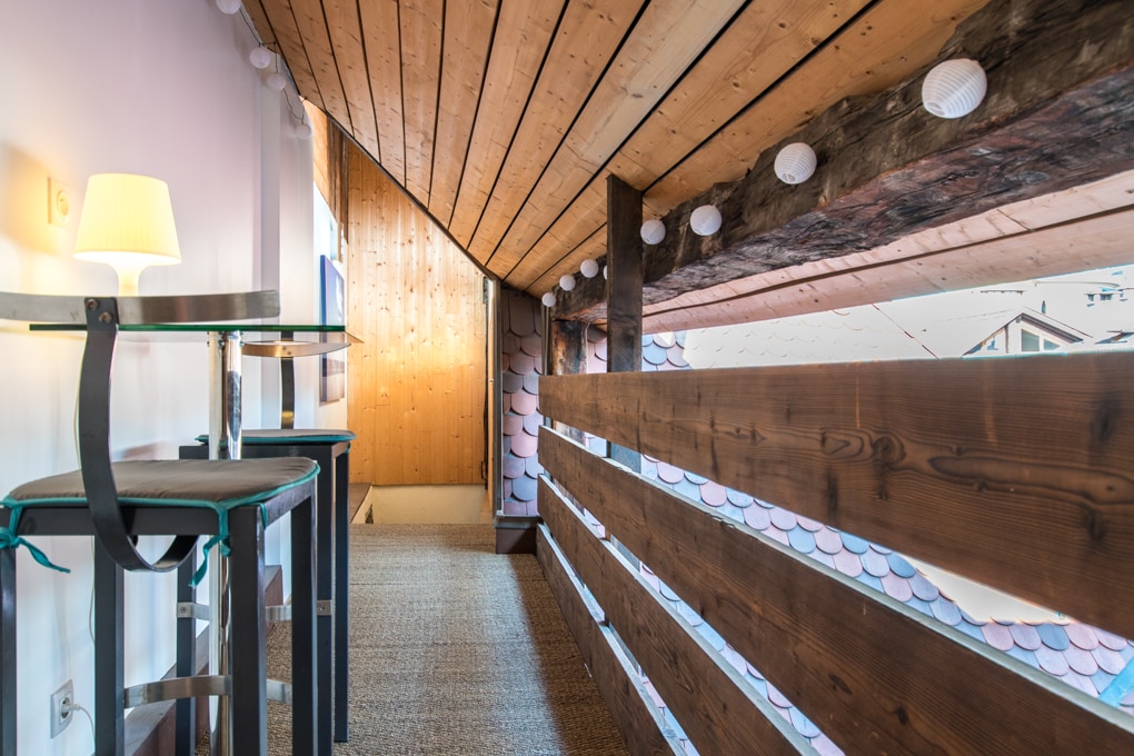 A hallway features wooden beams and a sloped ceiling, creating a cozy ambiance. A small table and a lamp are positioned on one side, while a wooden railing runs along the edge. Soft lighting is provided by decorative string lights overhead.