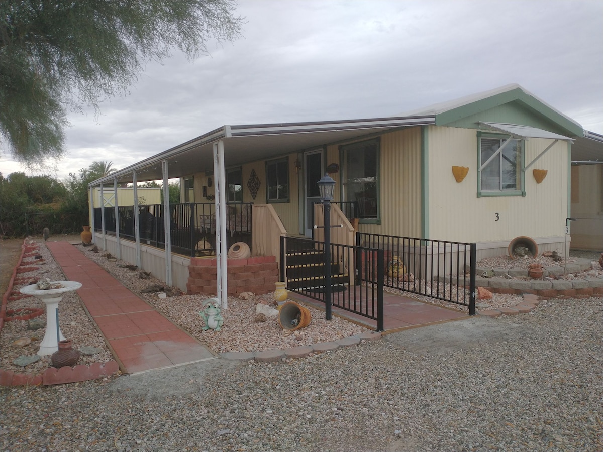 A welcoming entrance features a covered porch leading to the door. A pathway is lined with decorative elements, including stones and planters. The property is surrounded by gravel and a manicured garden, providing a serene outdoor space.