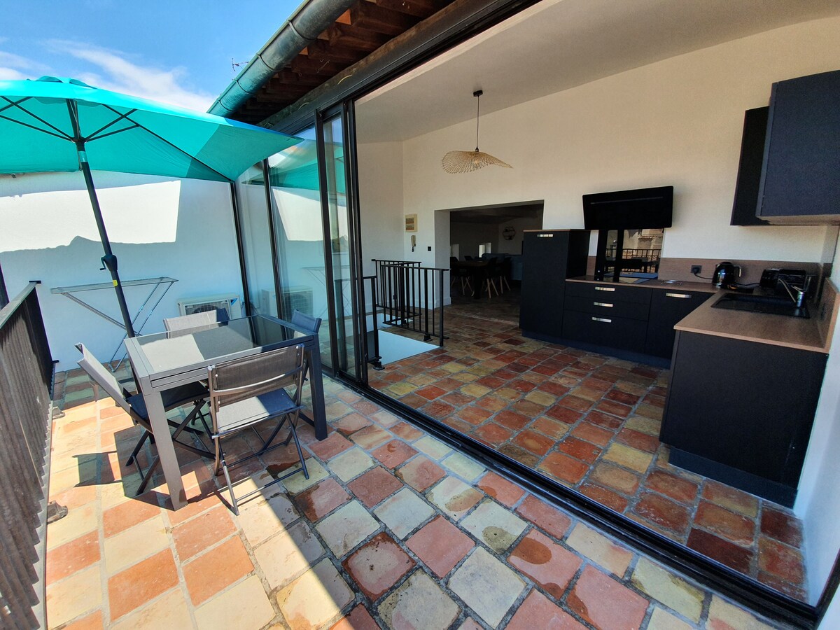 A terrace space is shown with stylish outdoor furniture, including a dining table and chairs. Large glass doors lead to an open interior that features a modern kitchen and living area. Sunshine highlights the terracotta tiled flooring and the vibrant turquoise umbrella.
