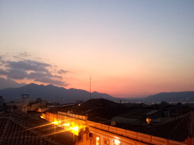 A serene sunset view is captured over the rooftops, with hues of orange and pink blending into the evening sky. The distant mountains create a soft silhouette against the colorful backdrop, while warm lights from nearby buildings illuminate the foreground.