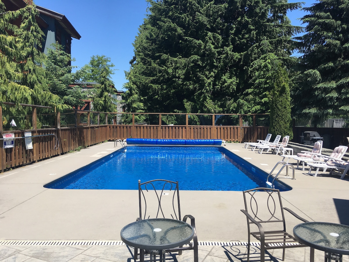 An outdoor swimming pool is situated within a fenced area, surrounded by sun loungers and tall trees. The pool features a clear, blue surface, reflecting the bright sky above. Concrete decking provides space for relaxation, while an inflatable pool cover rests along one edge.