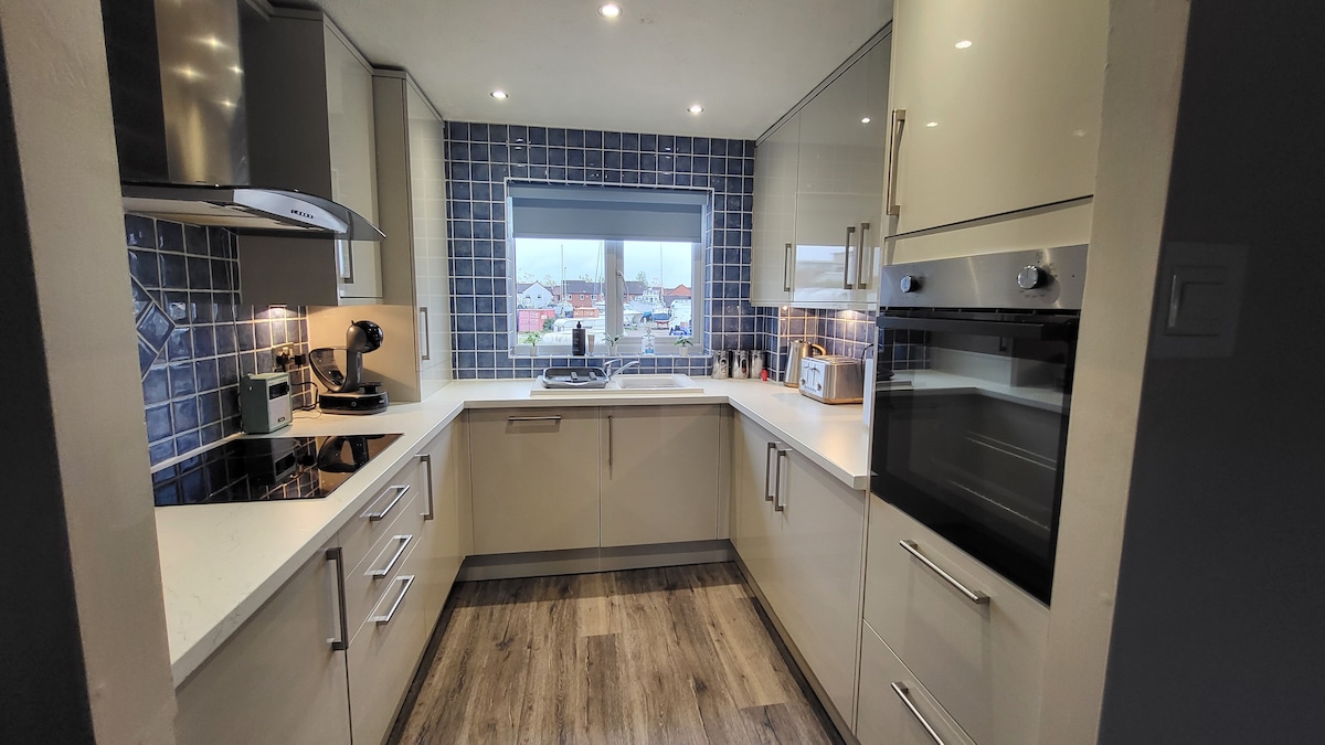 A well-equipped kitchen features modern cabinetry and a spacious countertop. Bright blue tiled backsplash complements the sleek design. Large windows offer natural light and a view of the surroundings. Appliances include an oven, microwave, and coffee machine, ensuring functionality for meal preparations.