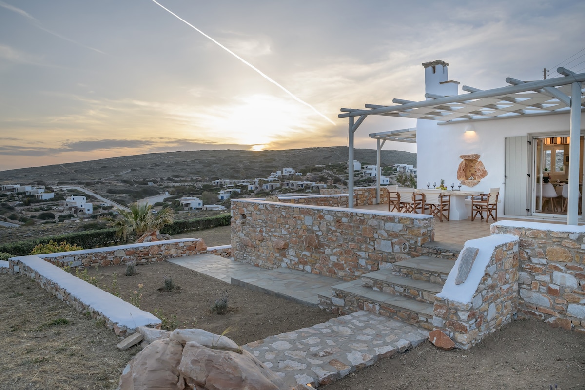 The exterior of a traditional Cycladic house is depicted, showcasing a stone wall and a terrace area. The sun sets over distant hills, casting a warm glow. Simple outdoor furniture is arranged on the patio, inviting relaxation and enjoying the surrounding landscape.