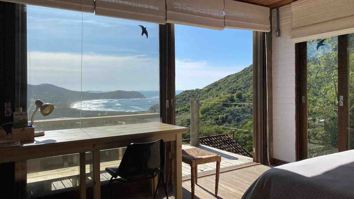 A spacious room featuring large glass windows that offer a panoramic view of the lush green hills and coastline. A sturdy wooden desk and a simple chair are positioned near the window, allowing for relaxation while enjoying the scenery.