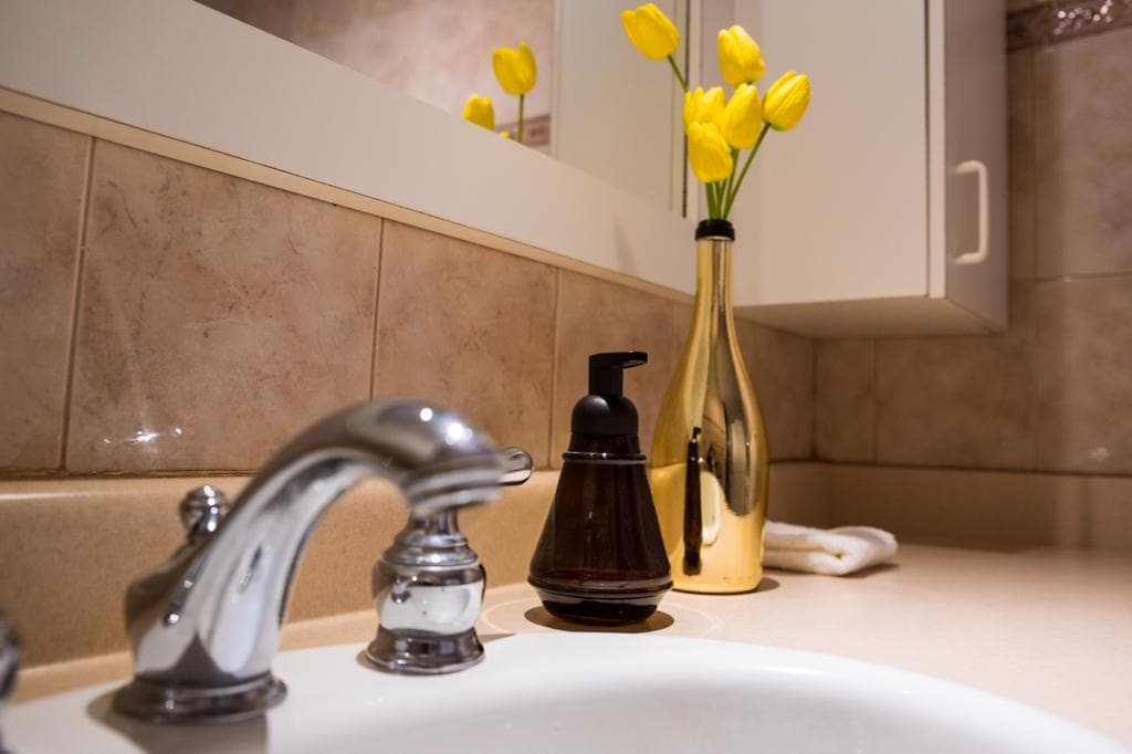 A bathroom sink features a polished chrome faucet and a dark-colored soap dispenser on the countertop. A golden vase holds vibrant yellow tulips, adding a touch of color against the soft-toned tiles of the wall and countertop.