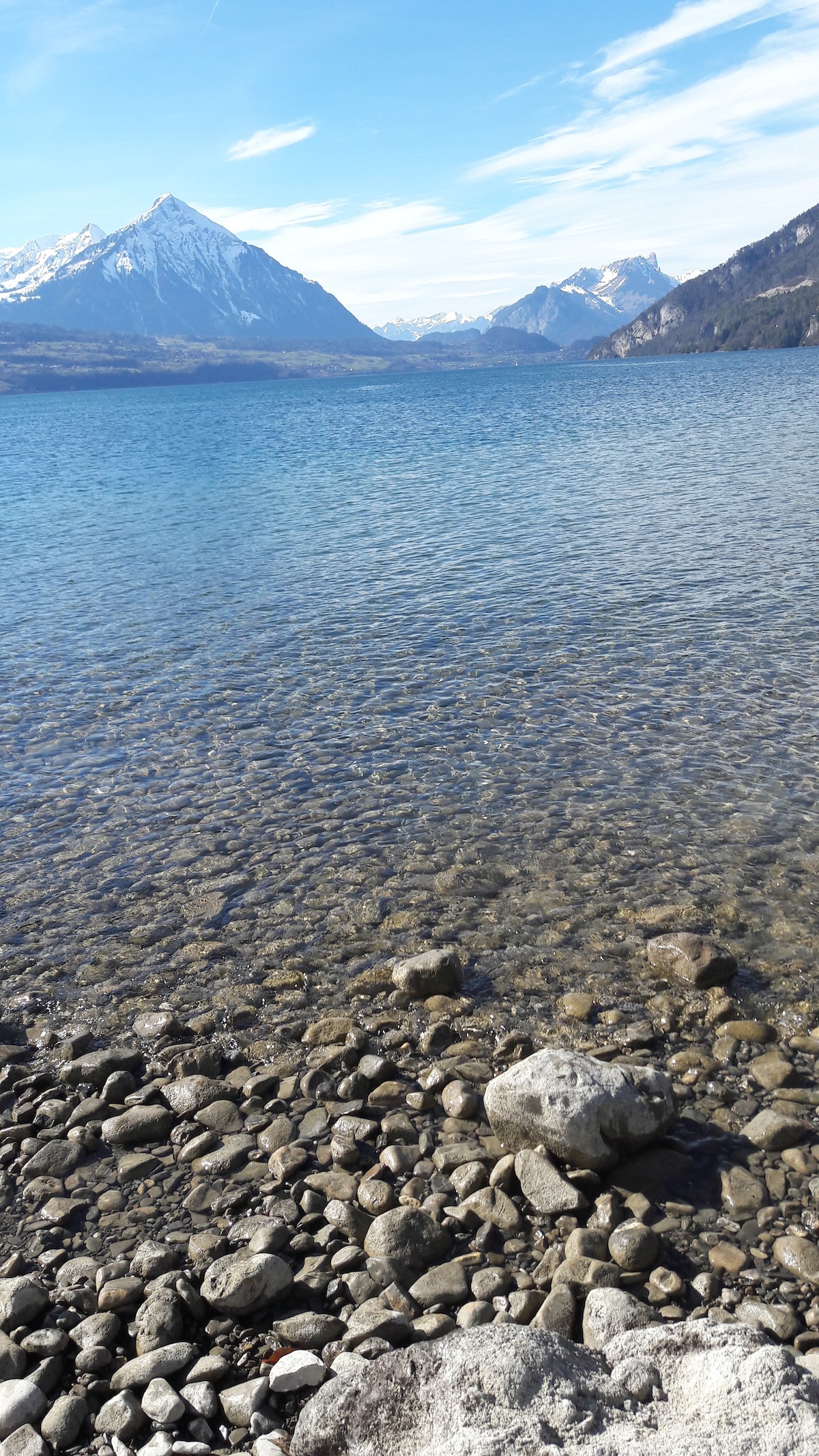 A calm lake scene presents clear blue waters reflecting the mountains in the background. Smooth stones line the water's edge, with various sizes and shapes visible. Snow-capped peaks rise majestically beyond the lake, under a bright sky with scattered clouds.