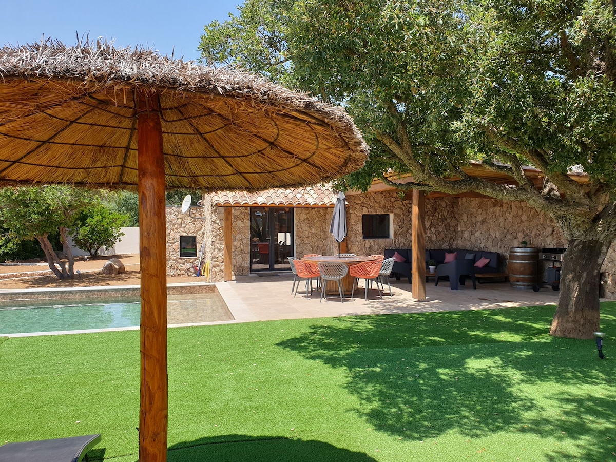 A shaded area features a thatched umbrella, offering a cool retreat beside a sparkling pool. The outdoor space showcases a dining table surrounded by seating and is enhanced by natural greenery. The stone structure blends harmoniously with the surrounding landscape.