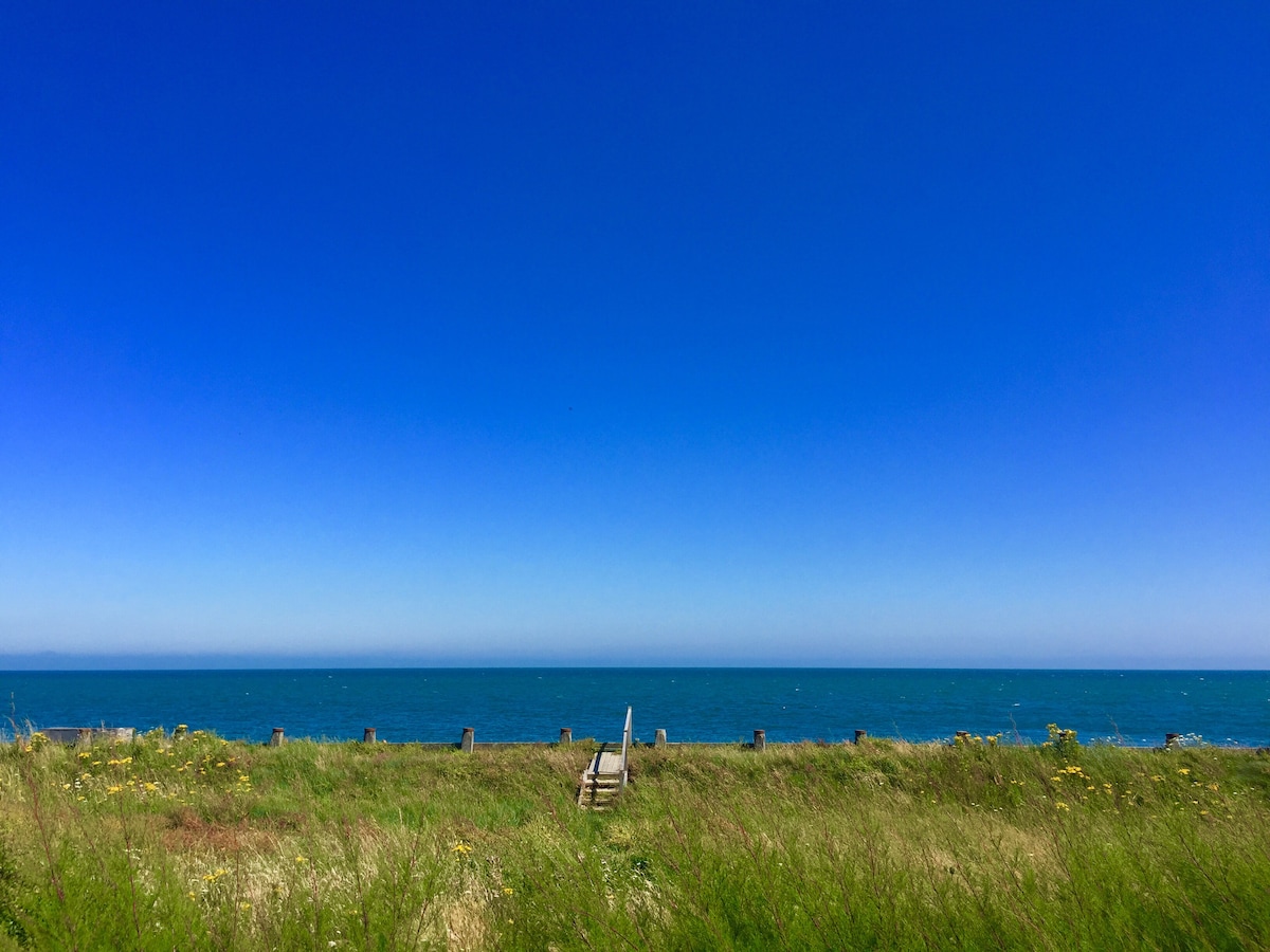 A serene coastal view features gentle waves meeting a clear blue sky. Lush green grass and wildflowers frame the scene, leading to a simple wooden beach access step at the shoreline.