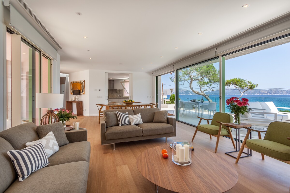 A bright living area features large glass doors that open to a scenic ocean view. Modern furniture includes a pair of armchairs and a spacious sofa adorned with neutral accents. A low coffee table holds a decorative arrangement, while a dining area is visible in the background.