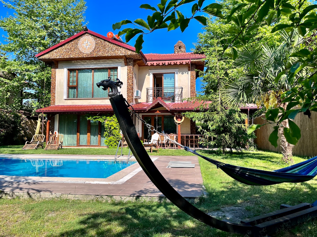 The exterior of the villa features a well-maintained garden with a private swimming pool and a hammock. A double-story structure is adorned with wooden accents. Large windows reflect natural light, while the sunny blue sky enhances the inviting ambiance.