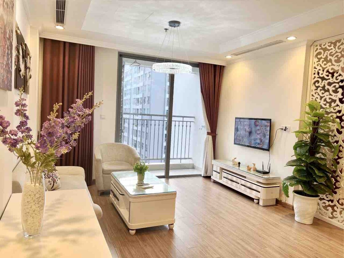A bright living area features a comfortable seating arrangement with a neutral color palette. Large windows allow natural light to enter, providing views of the balcony. A decorative table sits in the center, accompanied by potted plants and wall art that enhance the space.