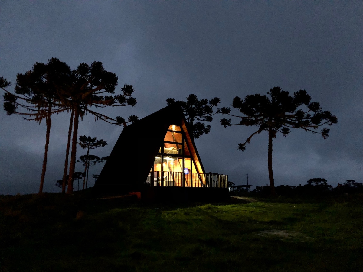 The chalet's unique A-frame structure is illuminated from within, standing amidst a dark landscape. Tall trees frame the scene, while the surrounding grass is softly lit by the building's glow, creating a contrasting visual against the evening sky.