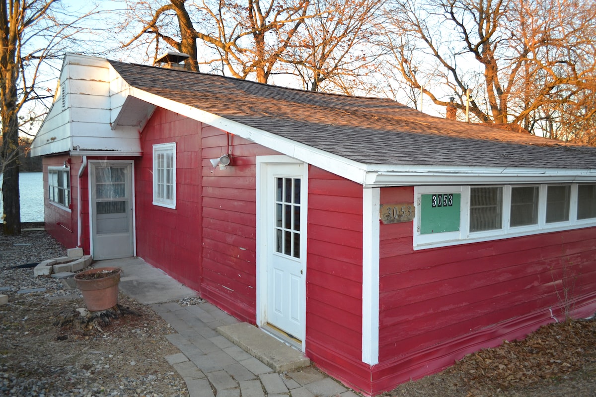 The exterior of Paradise Cottage features vibrant red wood paneling, with a stone path leading to a welcoming front door. Large windows allow natural light and views of the surrounding trees, enhancing the cottage's connection to its lakeside environment.