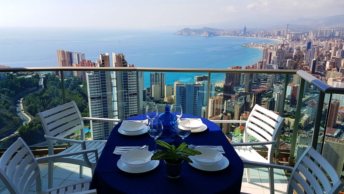 An outdoor dining table is set for four, adorned with white plates and glassware. The table is positioned on a balcony with a glass railing, offering unobstructed views of the sea and city of Benidorm. A small plant is placed in the center of the table.