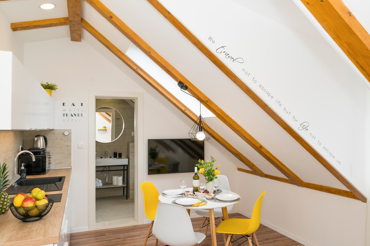 A bright dining area is showcased, featuring a round table set for four. Two yellow chairs complement the space, while natural light filters through the slanted roof. Modern kitchen appliances are visible, including a coffee maker and a bowl of fresh fruit on the counter.