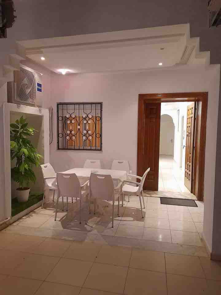 A bright dining area features a white table surrounded by six matching chairs, creating a clean and modern aesthetic. A green plant adds a touch of nature, while a decorative wall piece enhances the space. The area is well-lit with recessed ceiling lights.