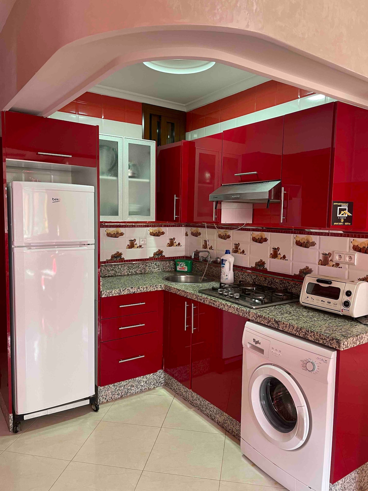A modern kitchen features rich red cabinetry complemented by granite countertops. Essential appliances include a refrigerator, gas stove, and washing machine. The kitchen layout is functional, with ample storage and illustrated tiles adorning the backsplash, creating a vibrant cooking space.