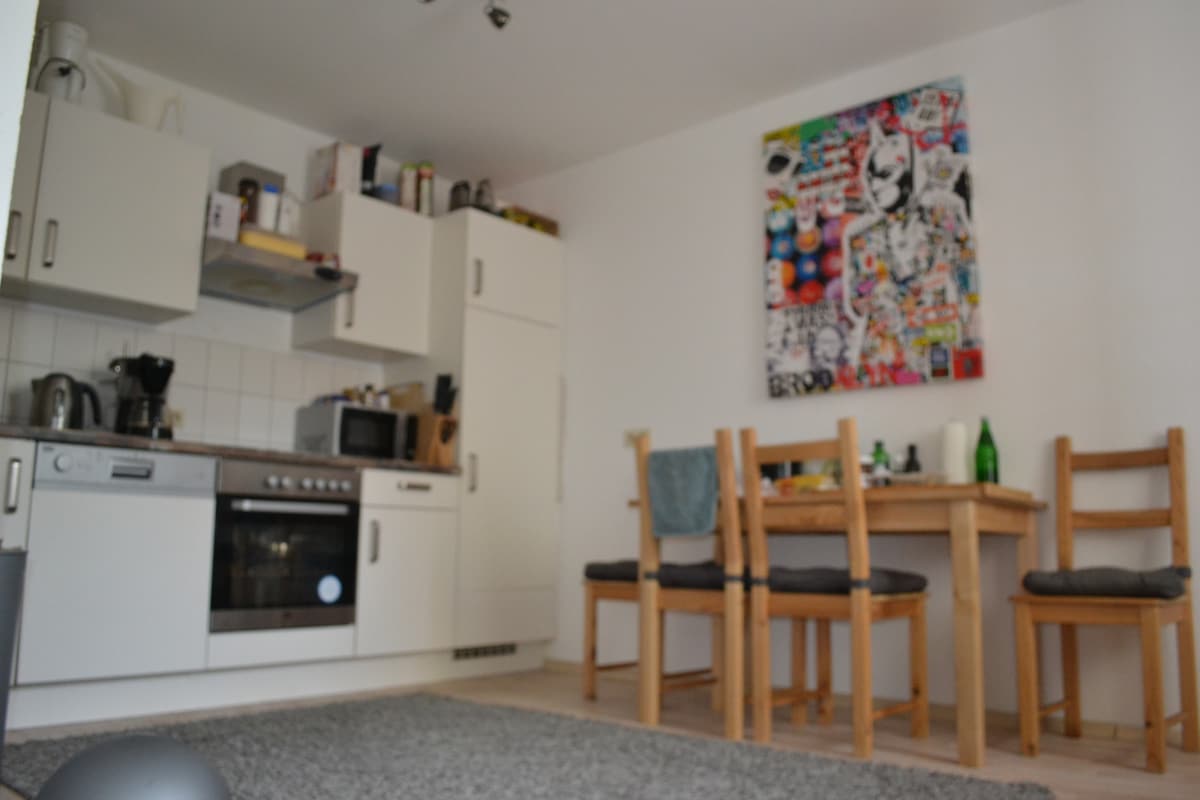 A modern kitchen area is depicted with sleek white cabinetry and a mirrored oven. The dining table is accompanied by four wooden chairs, while a colorful wall art piece adds visual interest. Kitchen appliances, including a kettle and coffee machine, are neatly arranged on the counter.