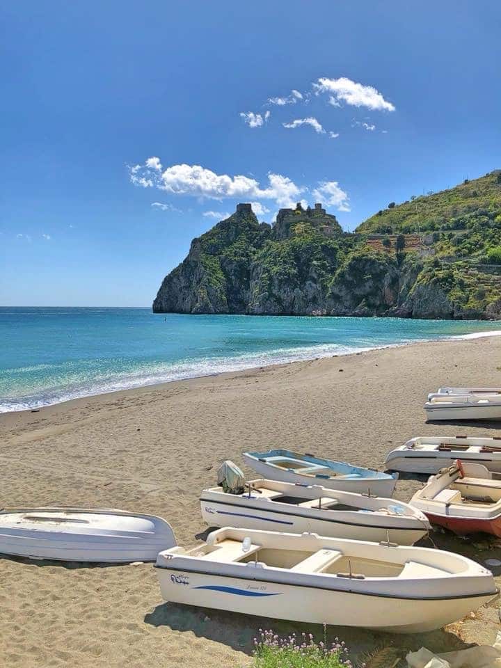 A serene beach scene is depicted with small boats resting on soft sand. The turquoise waves gently lap at the shoreline, while rugged cliffs rise in the background, creating a striking contrast against the clear blue sky adorned with wispy clouds.