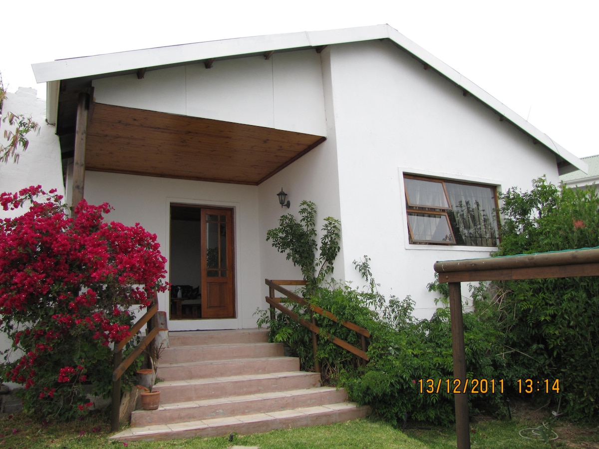 The exterior of the cottage is presented, showcasing a welcoming entrance with wooden double doors and steps leading up to the porch. Bougainvillea in vibrant pink climbs along the side, while greenery enhances the inviting appeal of the surrounding garden.