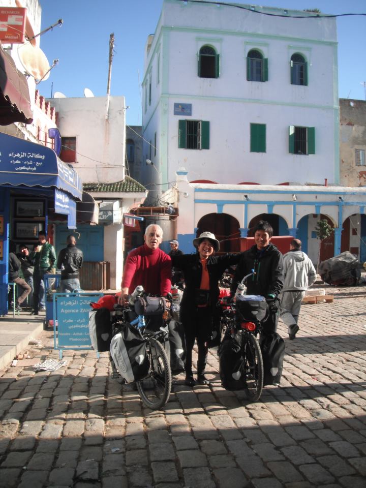 A vibrant street scene is captured, featuring three individuals standing beside bicycles equipped with bags. The background displays a historic building with blue and white walls, complemented by green shutters. Cobblestone surfaces add texture to the environment, reflecting the lively atmosphere of the medina.