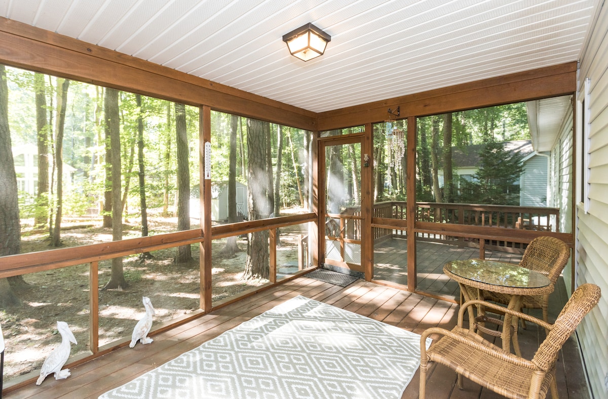 A screened-in porch offers views of the surrounding forest, featuring wooden beams and a ceiling light. A small round table with a glass top sits beside a wicker chair, positioned on a geometric-patterned rug. Two decorative rabbit figures are placed on the wooden railing.