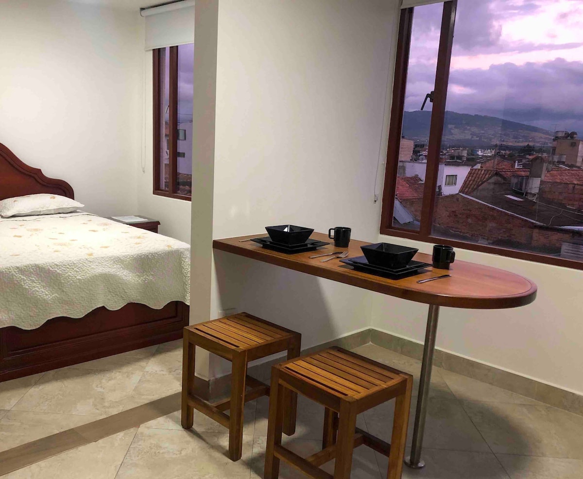 A spacious room features a comfortable double bed and a dining area with a wooden table and two matching stools. Large windows provide views of the surrounding landscape, allowing natural light to brighten the space.