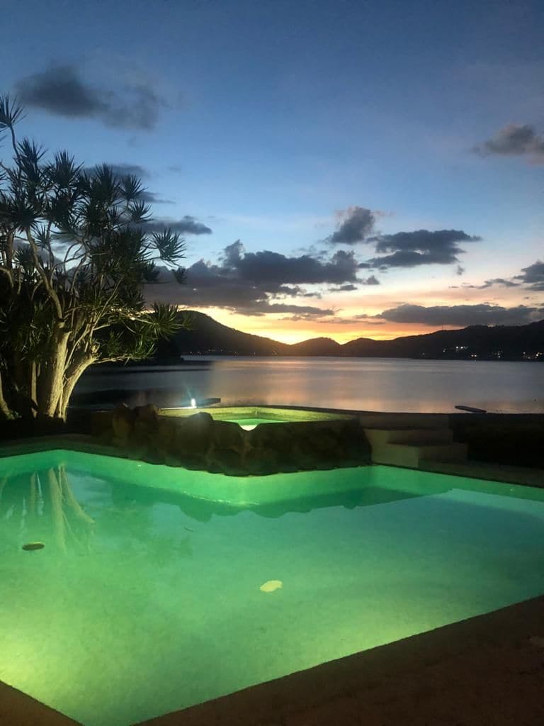 A tranquil pool reflects the colorful sunset over the lake, surrounded by lush greenery. Subtle lighting enhances the serene atmosphere, and distant mountains create a picturesque backdrop.