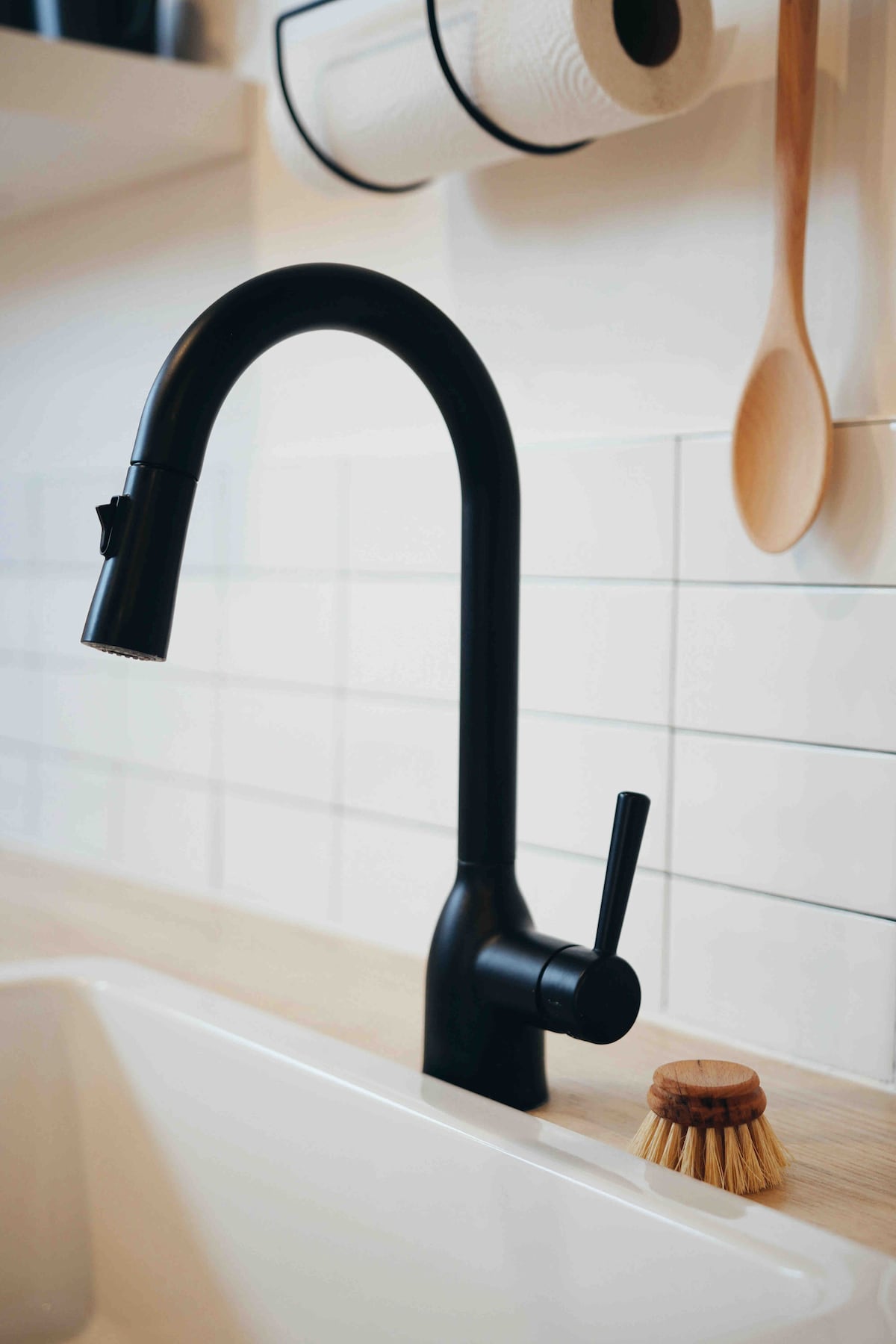 A sleek black kitchen faucet is positioned over a spacious white sink, featuring a curved spout and separate lever for temperature control. Nearby, a natural bristle scrub brush rests on the countertop, contrasting with the clean, minimalist design of the surroundings.