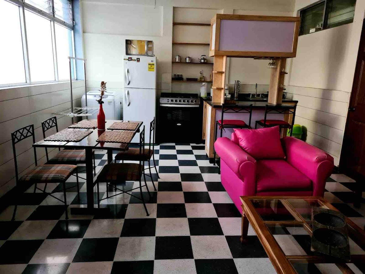 The open kitchen and dining area is complemented by checkered flooring. A glass dining table with four chairs sits adjacent to a pink sofa and a coffee table. The kitchen includes a refrigerator, stove, and various kitchen utensils, with natural light coming through large windows.