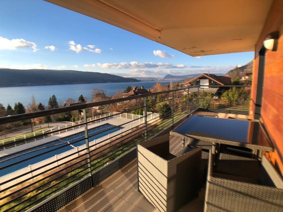 A private terrace is shown featuring outdoor seating with a table and chairs. The panoramic view includes the lake and surrounding landscape with a swimming pool visible below. The terrace is bathed in natural light, creating a welcoming outdoor space.