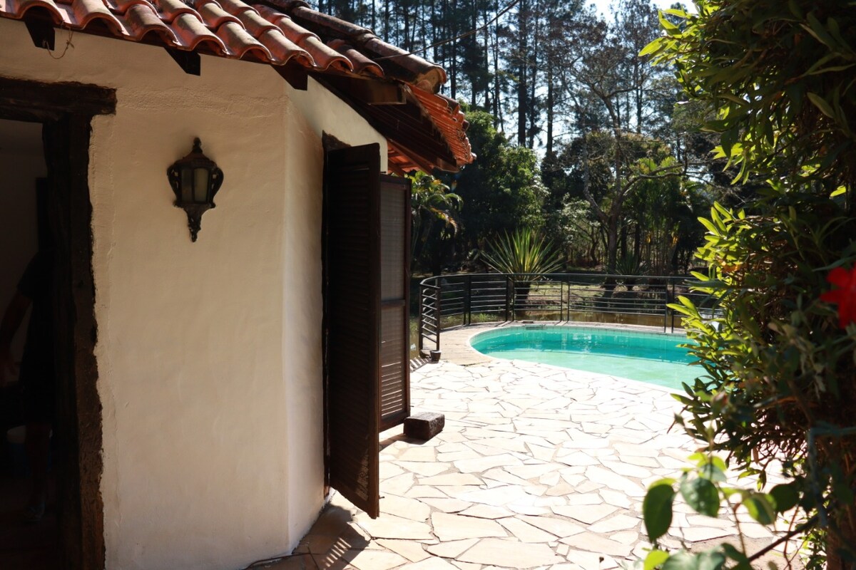 An exterior view showcases a charming structure with a tiled roof, revealing open wooden shutters. A stone pathway leads to a serene swimming pool, surrounded by lush greenery and trees in the background, inviting relaxation in a natural setting.