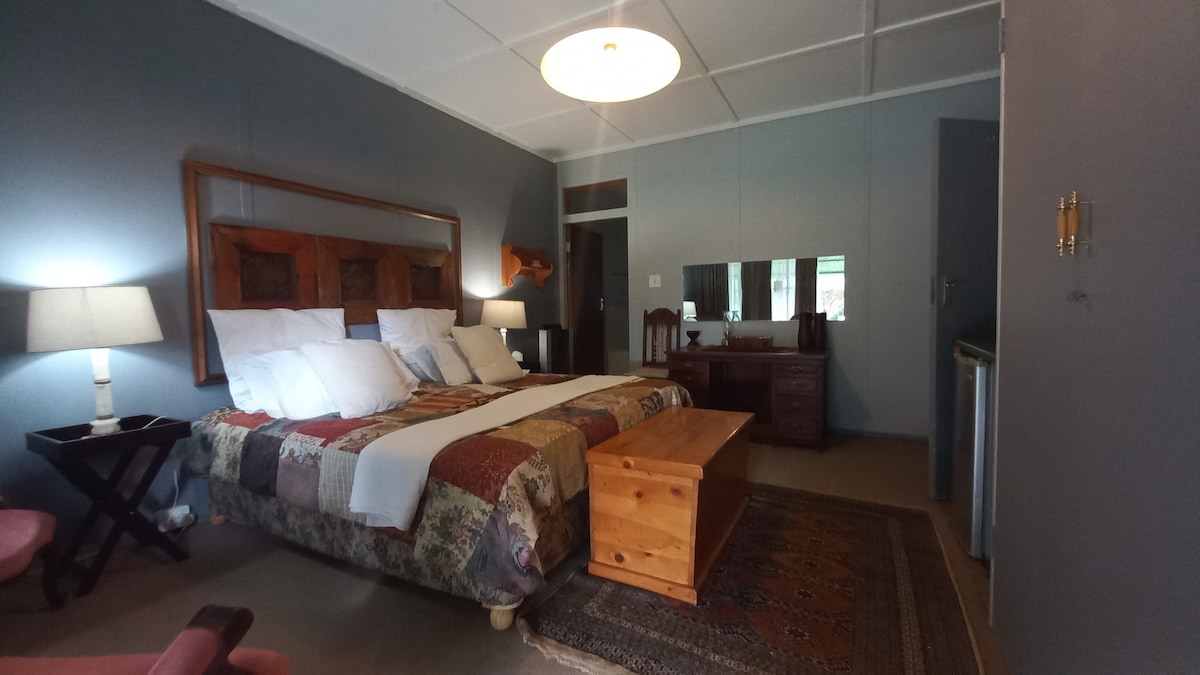 A well-furnished bedroom features a spacious bed with decorative bedding, complemented by bedside lamps and a wooden table. A mirrored dresser and mini fridge are visible. Neutral walls and a patterned rug enhance the room's inviting ambiance.