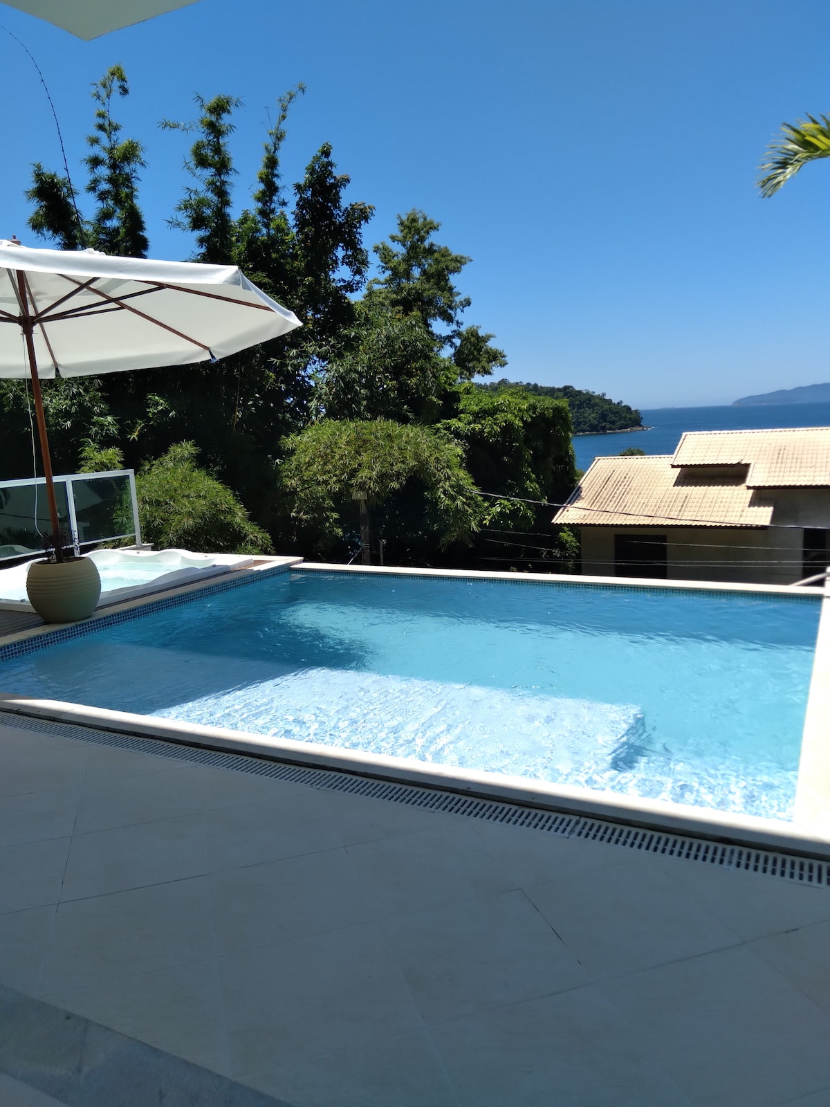 A clear swimming pool is situated at the edge of a terrace, surrounded by lush greenery. A large umbrella provides shade, and the view extends toward distant hills and the ocean. The bright blue sky enhances the serene atmosphere of the outdoor space.