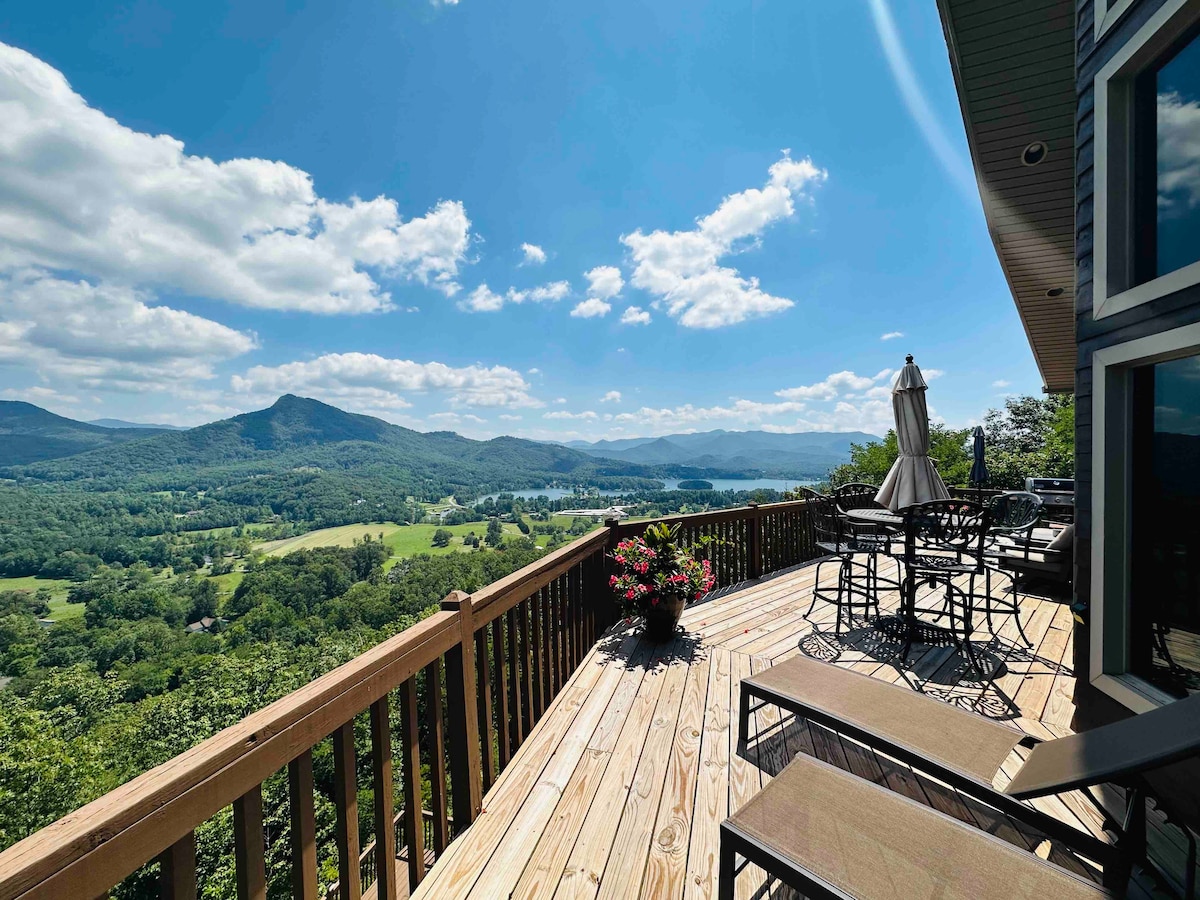 A spacious deck overlooks breathtaking views of rolling hills and Lake Chatuge. Several patio chairs and a dining table are arranged on the wooden surface, while potted flowers add a touch of color. Sunlight bathes the area, highlighting the panoramic landscape.