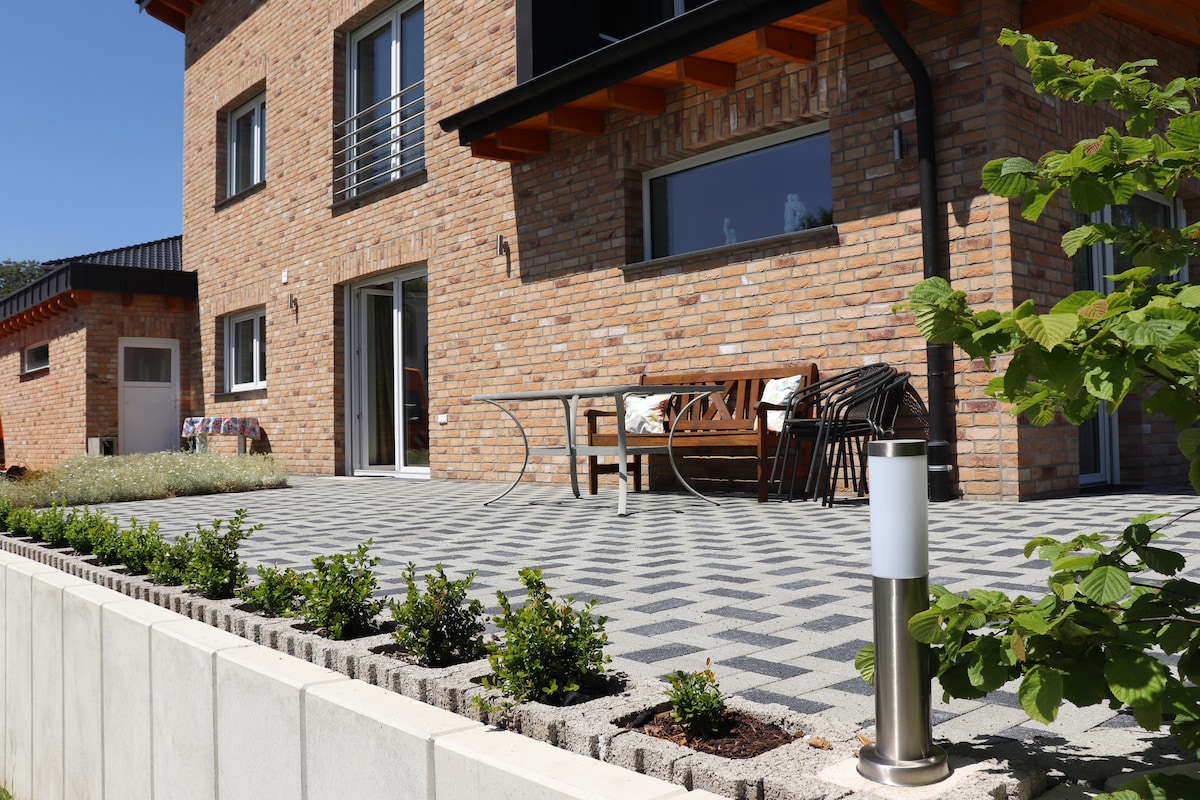 A spacious outdoor terrace features a wooden dining table surrounded by chairs. Neatly arranged greenery and small shrubs line the edge of the patio, while a tall, modern light fixture provides illumination. The brick façade of the house complements the serene atmosphere.