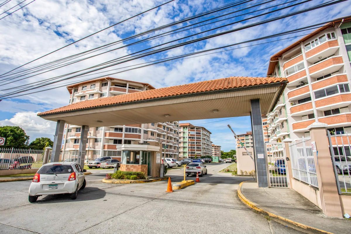 An entrance gate is visible, leading into a residential community. The structure features a canopy supported by columns, and vehicles are seen driving in. Several multi-story buildings with balconies line the approach, with blue skies and scattered clouds in the background.