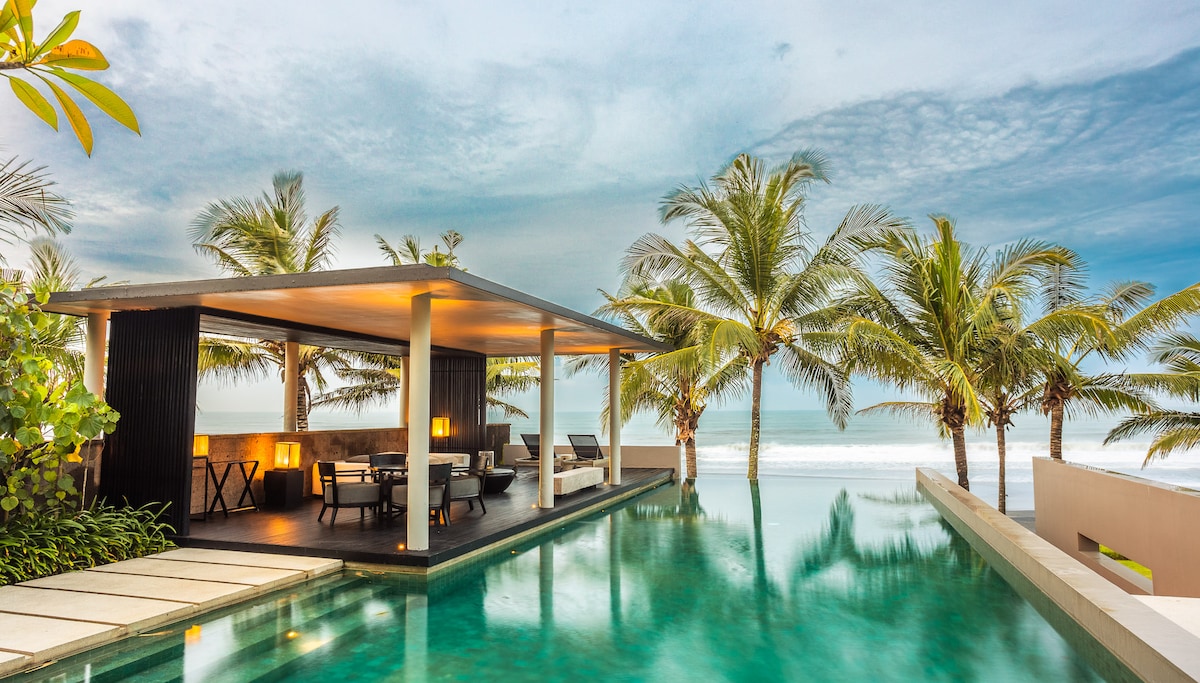 An open-air pavilion overlooks a large infinity pool, surrounded by tropical palm trees. The seating area features stylish outdoor furniture, creating a serene space for relaxation. The ocean is visible in the background, complementing the tranquil setting.
