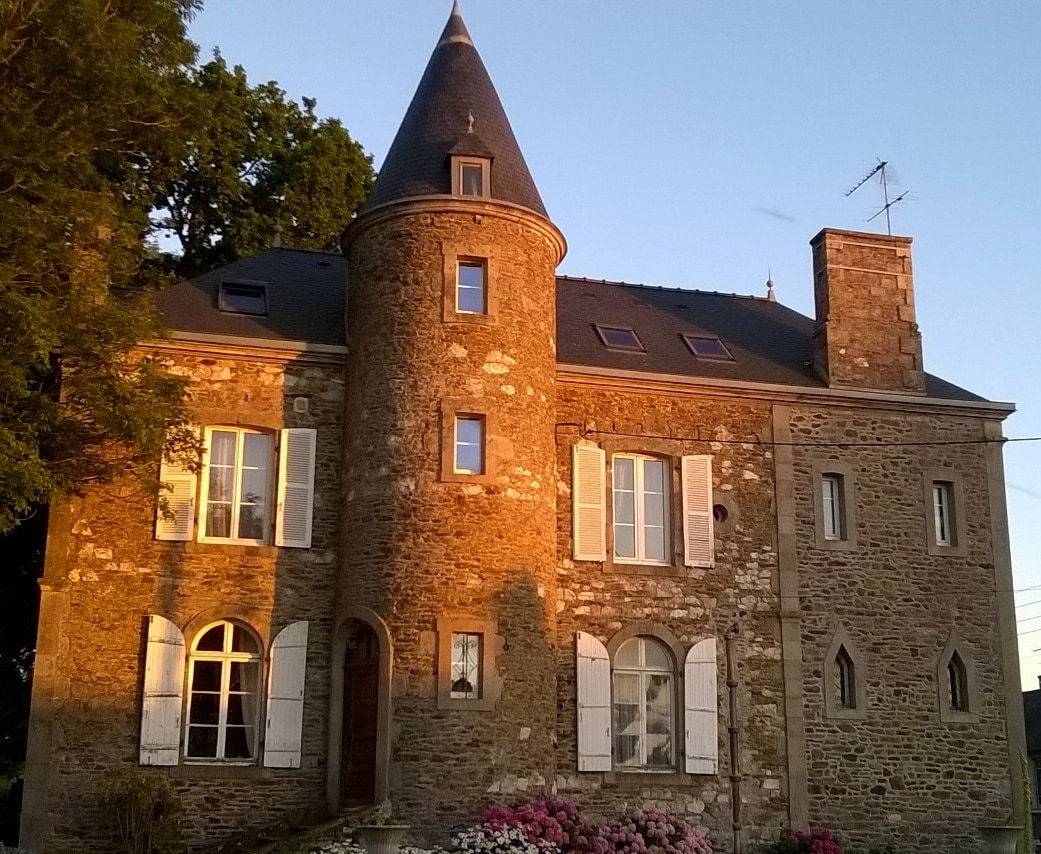 The 19th-century manor showcases a stunning stone facade with a rounded turret. Graceful windows are framed by white shutters, and vibrant pink flowers are seen in the foreground, adding color to the setting. Soft sunlight highlights the texture of the stone exterior.