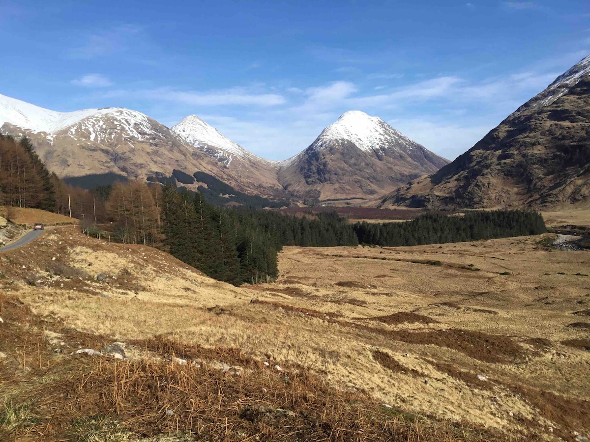 A panoramic view of the stunning Glen Etive landscape showcases the towering snow-capped mountains flanking the valley. The foreground features expansive, dry grassland, while dense patches of trees are visible at the base of the mountains. A clear blue sky enhances the serene atmosphere.