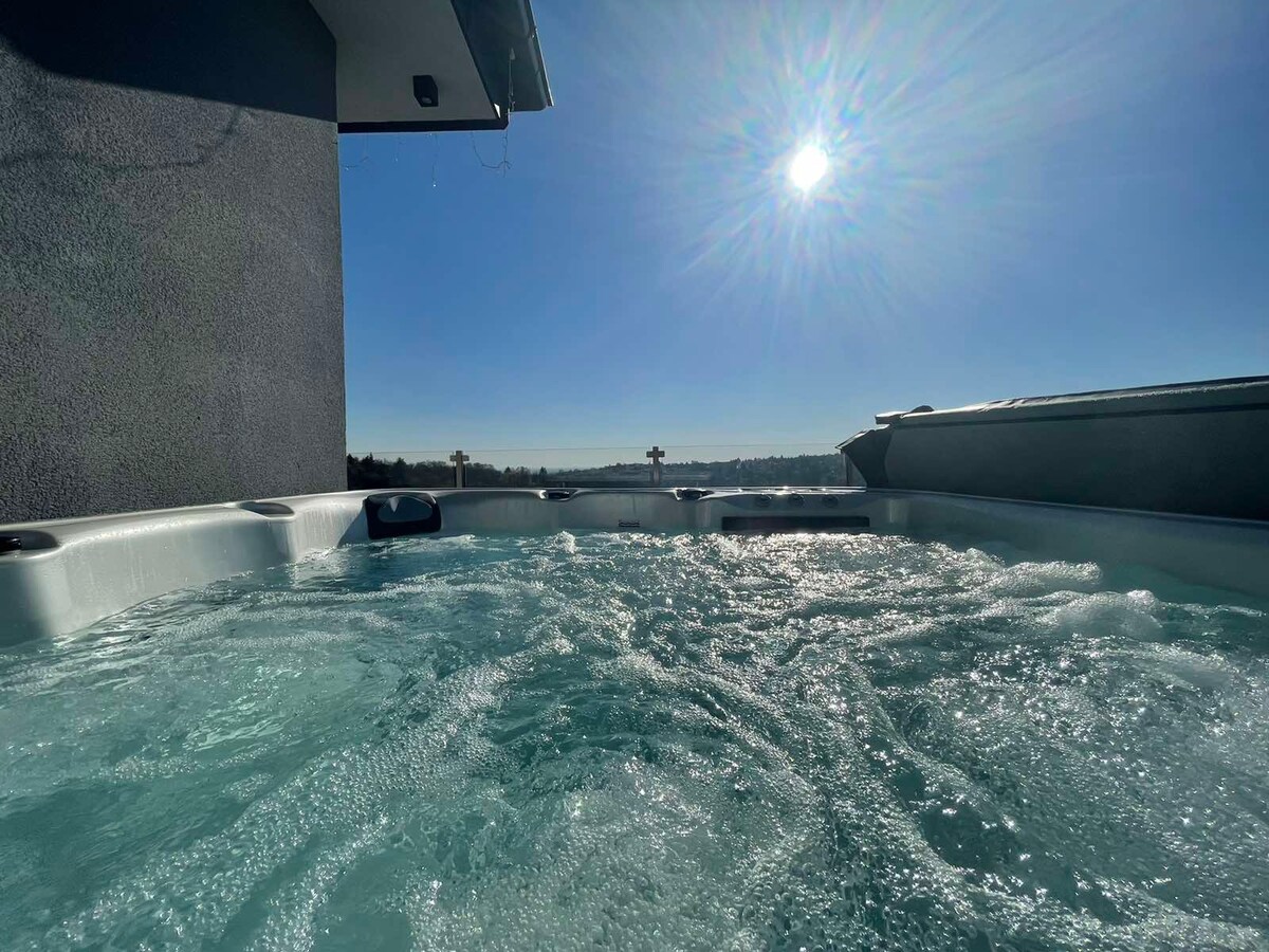 A view of the hot tub, filled with bubbling water, under a clear blue sky. Sunlight reflects off the surface, creating a bright atmosphere. Scenic vistas are visible in the background, enhancing the relaxing environment of the rooftop area.