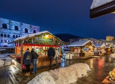 A winter market scene is displayed with wooden stalls adorned with colorful decorations. Snow-covered wooden pathways lead to various vendors, where people are seen browsing crafts and seasonal goods. A historical structure is illuminated in the background, enhancing the festive ambiance.