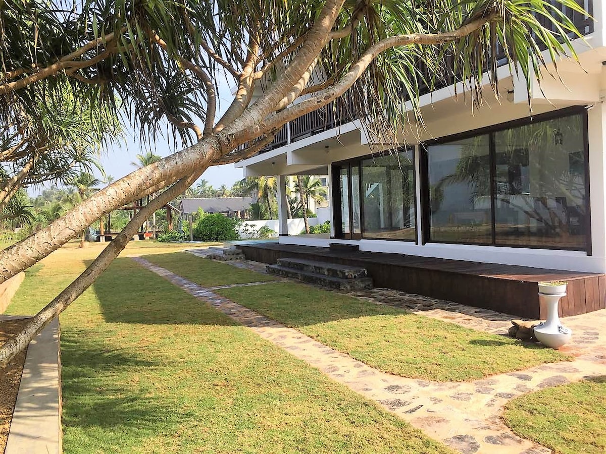 The exterior of the building features large glass windows that offer views of the surrounding garden. A well-maintained lawn leads to a wooden deck area, with stone pathways guiding through the greenery. Palm trees provide shade, contributing to a serene outdoor environment.