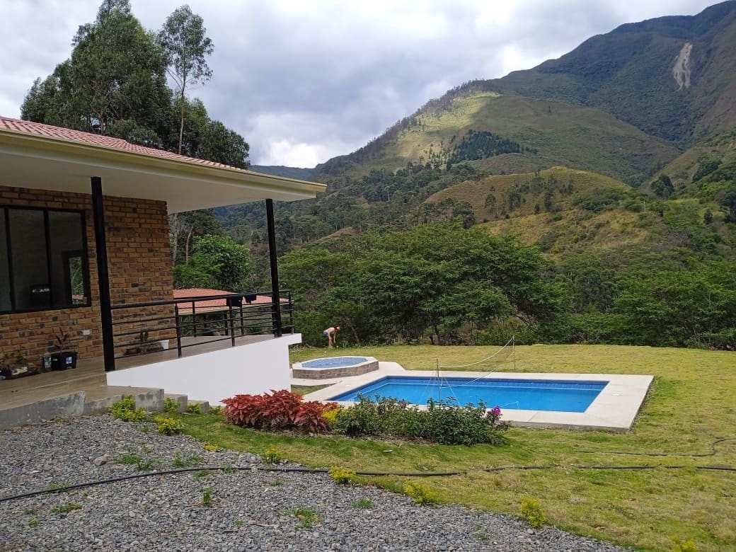 The image showcases a spacious outdoor area featuring a swimming pool and a jacuzzi, surrounded by verdant hills. A modern, brick house is positioned nearby, with large windows reflecting the natural landscape. The sky above is partly cloudy, casting gentle shadows on the ground.