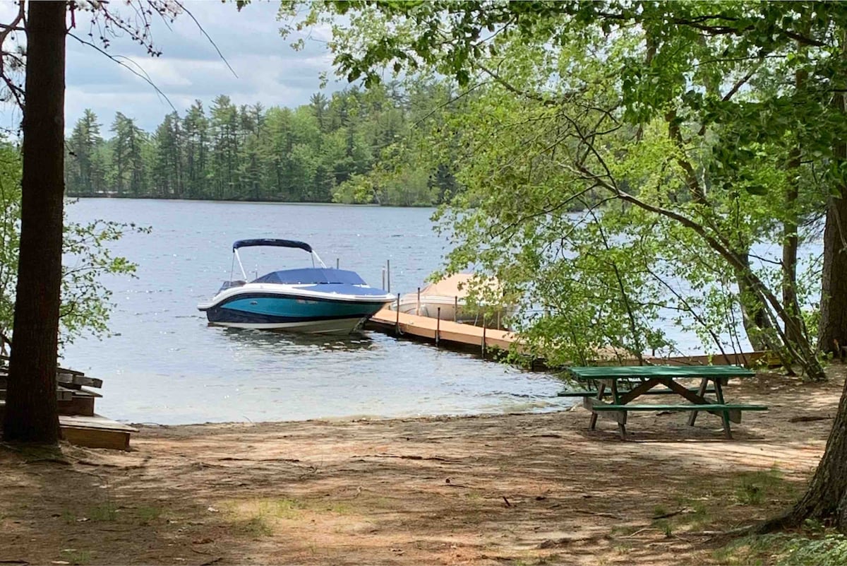A scenic view of a tranquil lake is captured, with a boat dock extending into the water. A boat is anchored near the dock, surrounded by lush greenery. A picnic table sits on sandy ground nearby, inviting relaxation by the lakeside.