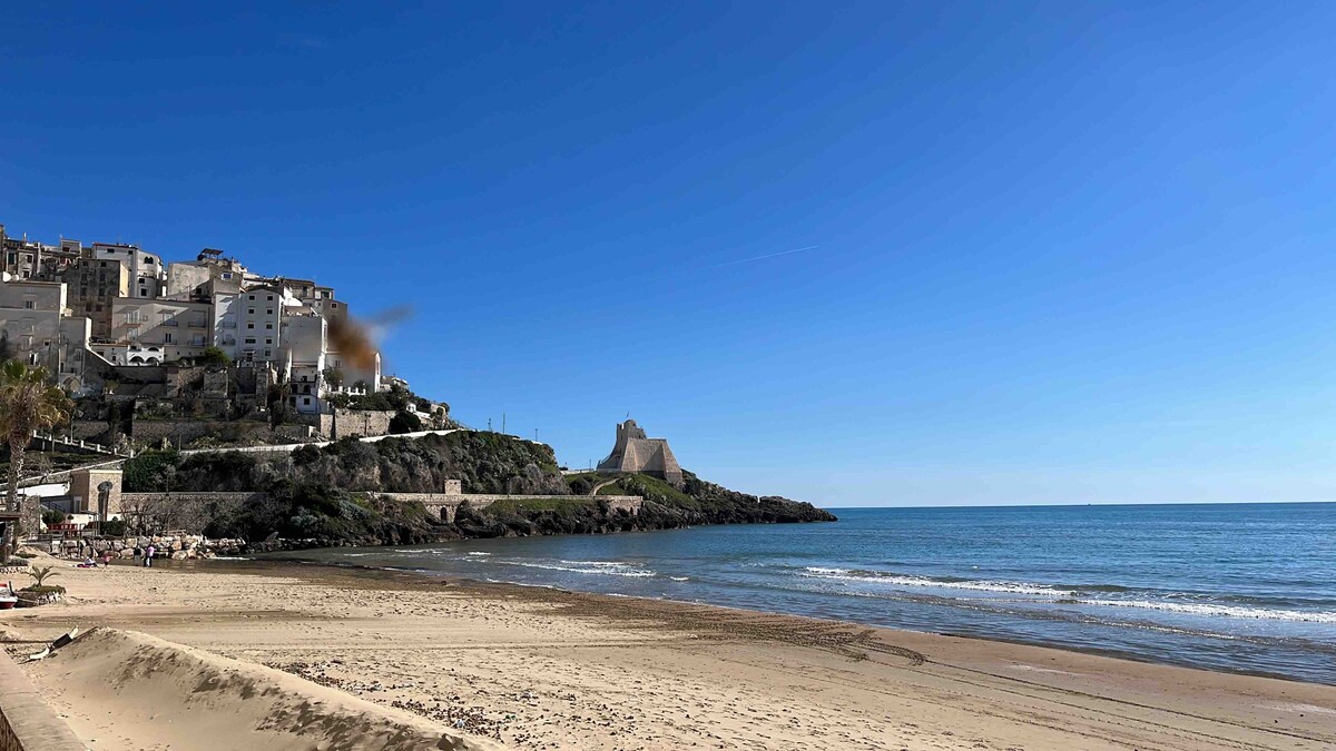 A scenic beach scene captures the shoreline with soft sand and gentle waves lapping at the shore. In the background, a historical tower and coastal village are visible, framed by a clear blue sky and sunlit waters.
