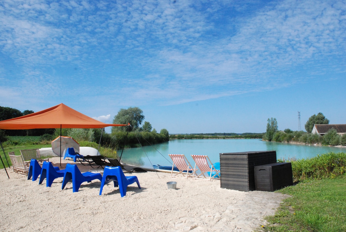 A sandy area alongside a calm lake is furnished with blue deck chairs and pink lounge chairs under an orange canopy. A wooden storage box is situated nearby, with greenery framing the water and a clear blue sky above.