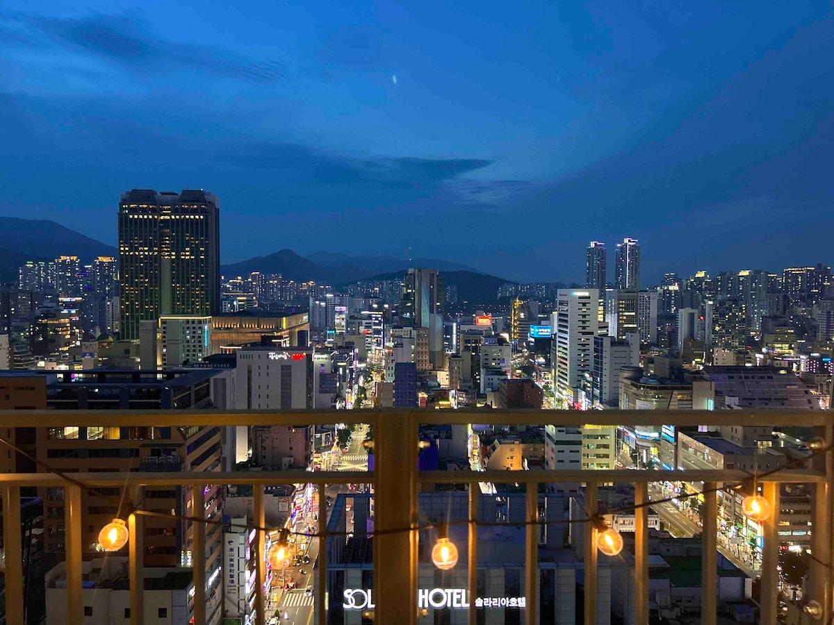 A panoramic city view is showcased from the balcony, with a vibrant skyline illuminated at night. Distant mountains provide a backdrop to the bustling streets below, while warm string lights enhance the outdoor space.