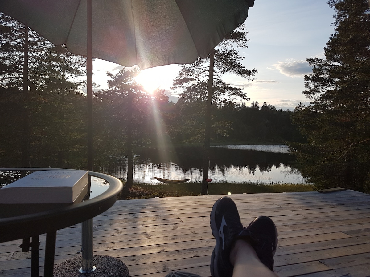 A scenic view of a tranquil lake is captured during sunset. A large umbrella provides shade for a circular table, while the wooden deck extends towards the water. A small boat is visible on the calm surface of the lake, surrounded by tall trees.