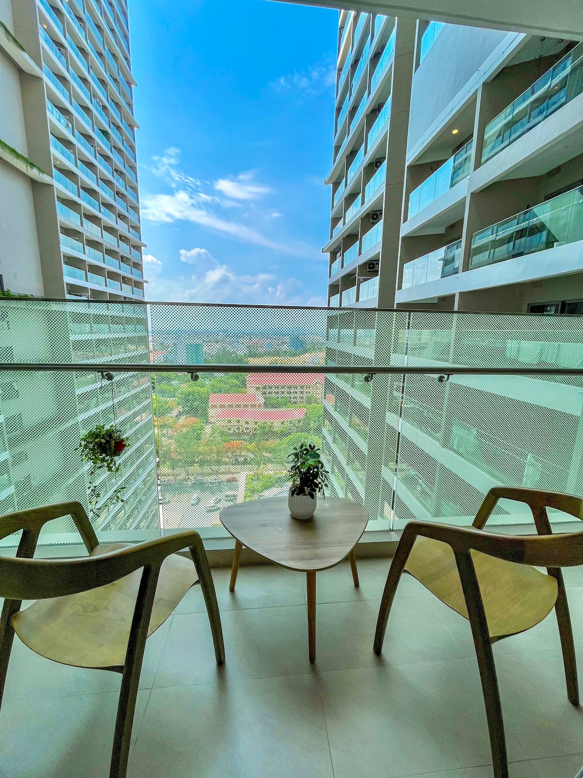 A balcony view is showcased with two wooden chairs positioned around a small table. Lush greenery is visible in planters, and the expansive cityscape is observed in the background under a clear blue sky.